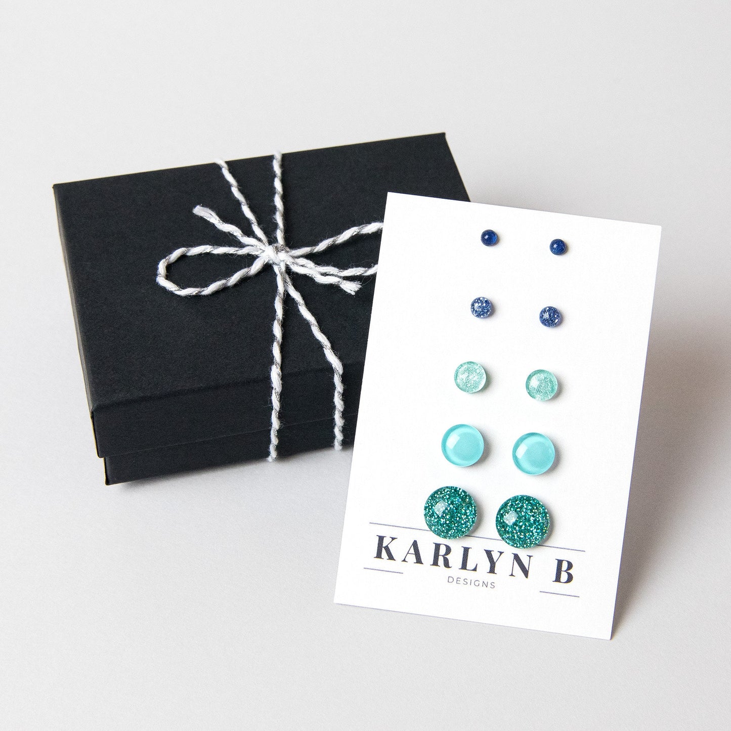 A set of five pairs of stud earrings in a black gift box. The earrings are in shades of blue and green, with some having glitter. The white earring card reads "KARLYN B DESIGNS".