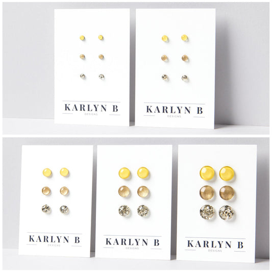 Five sets of stud earrings on white cards with the text "KARLYN B DESIGNS". Each set features three pairs of earrings in different shades of warm yellow, gold and gold glitter.