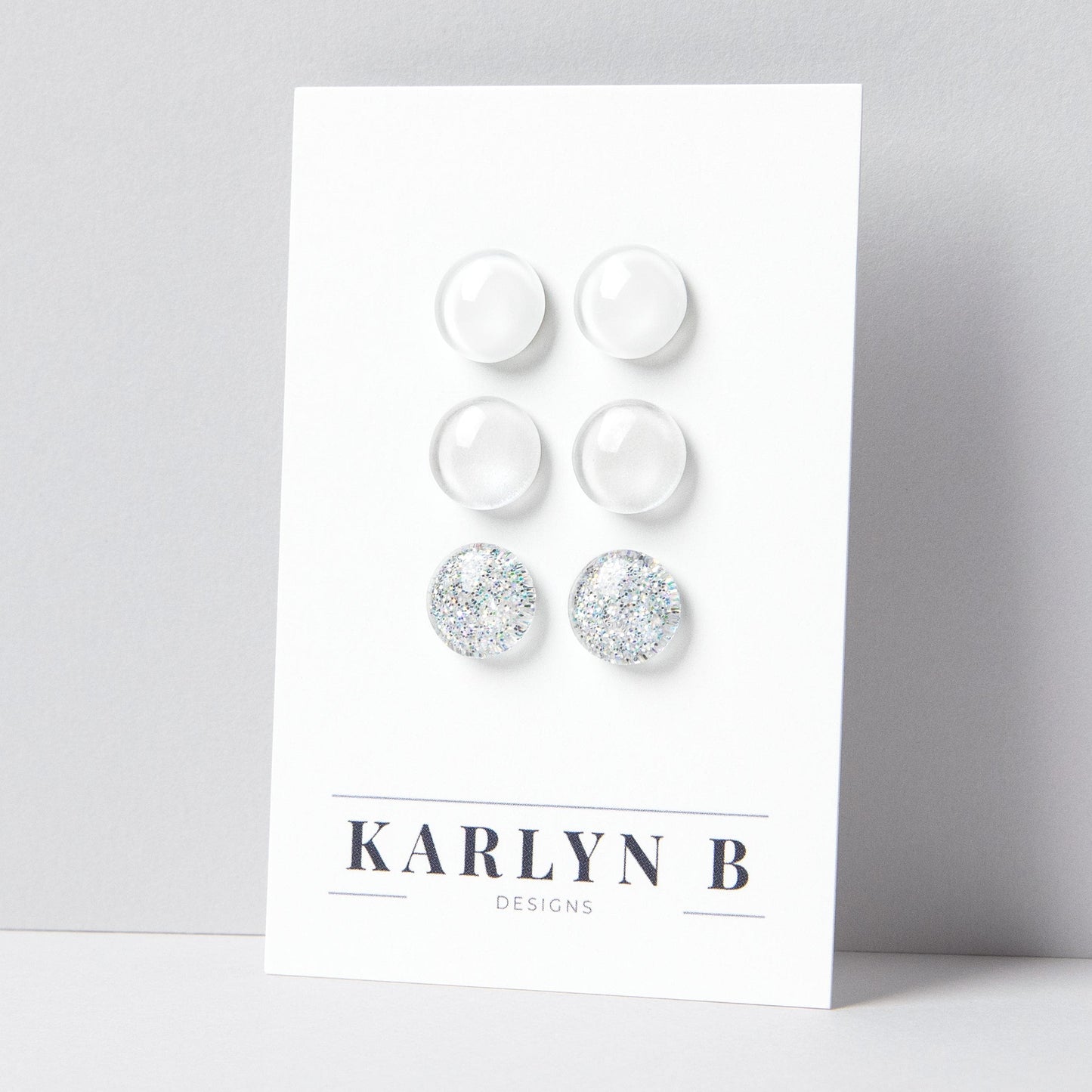 Set of three stud earrings in three different finishes - solid, shimmer, and glitter. The earrings are white solid, white shimmer, and white silver glitter. The earrings are displayed on a white card with the text "KARLYN B DESIGNS".