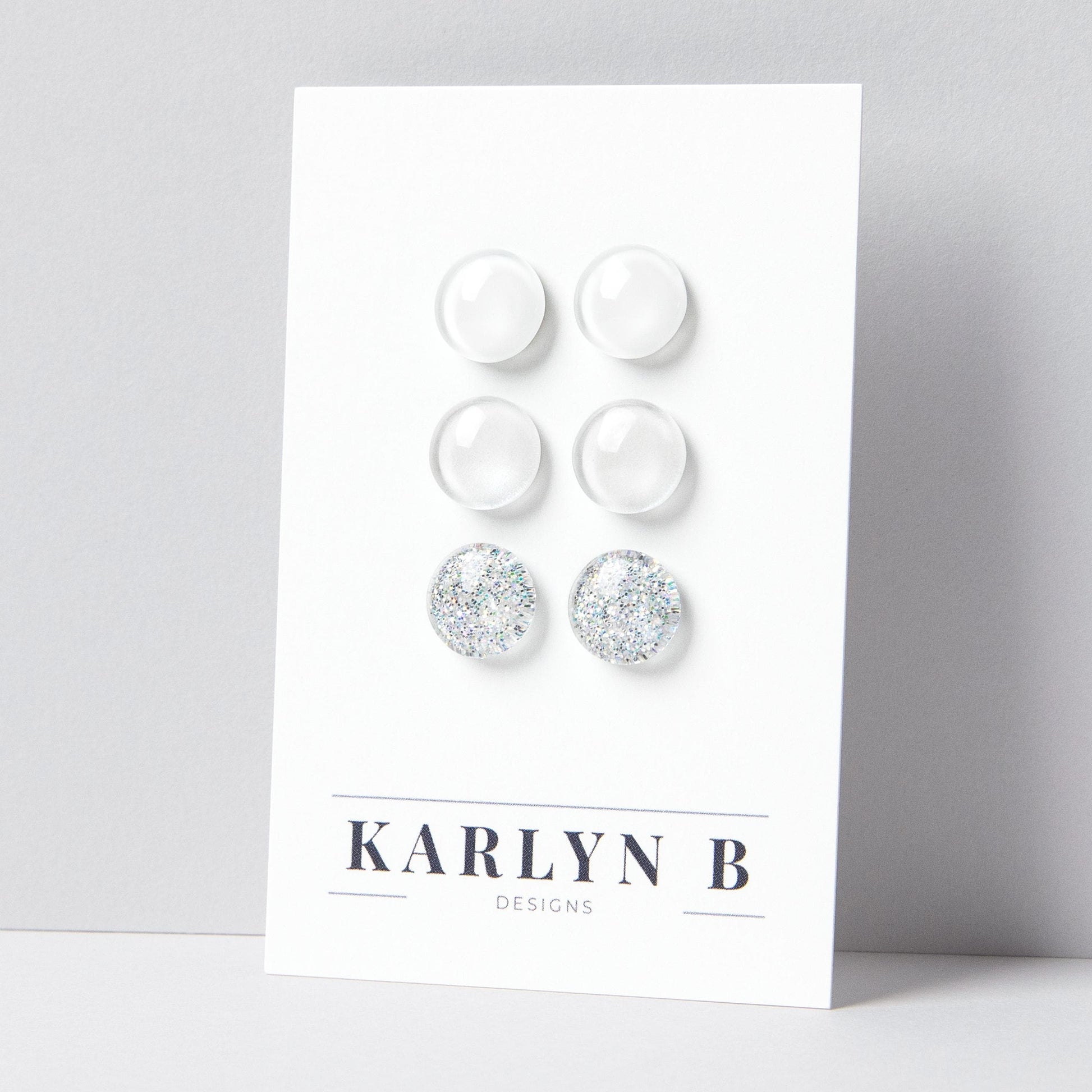 Set of three stud earrings in three different finishes - solid, shimmer, and glitter. The earrings are white solid, white shimmer, and white silver glitter. The earrings are displayed on a white card with the text "KARLYN B DESIGNS".