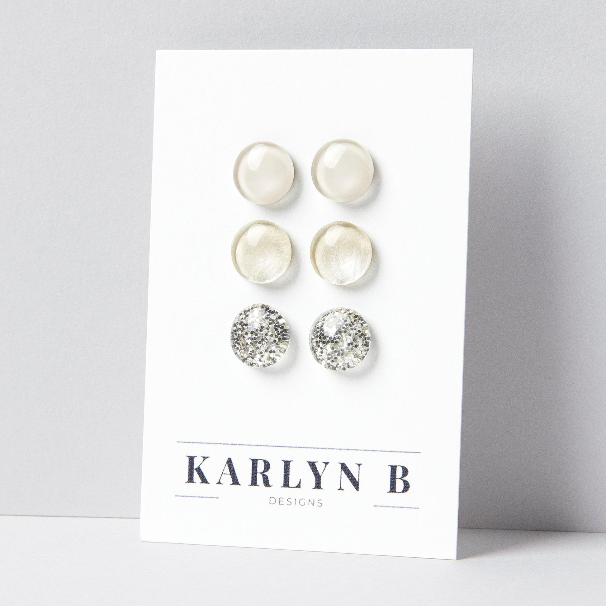 Set of three stud earrings in three different finishes - solid, shimmer, and glitter. The earrings are off white solid, ivory shimmer, and ivory silver glitter. The earrings are displayed on a white card with the text "KARLYN B DESIGNS".