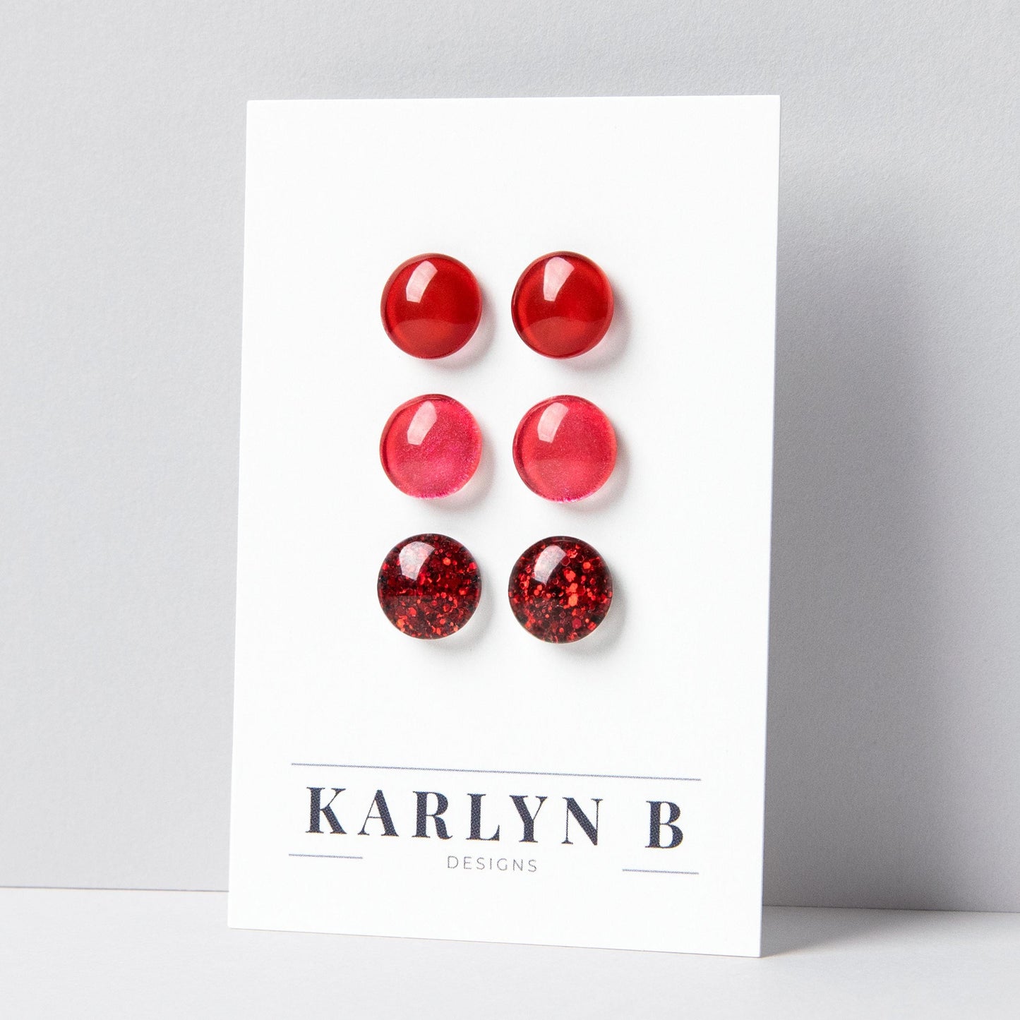 Set of three stud earrings in three different finishes - solid, shimmer, and glitter. The earrings are cherry red solid, ruby red shimmer, and ruby red glitter. The earrings are displayed on a white card with the text "KARLYN B DESIGNS".