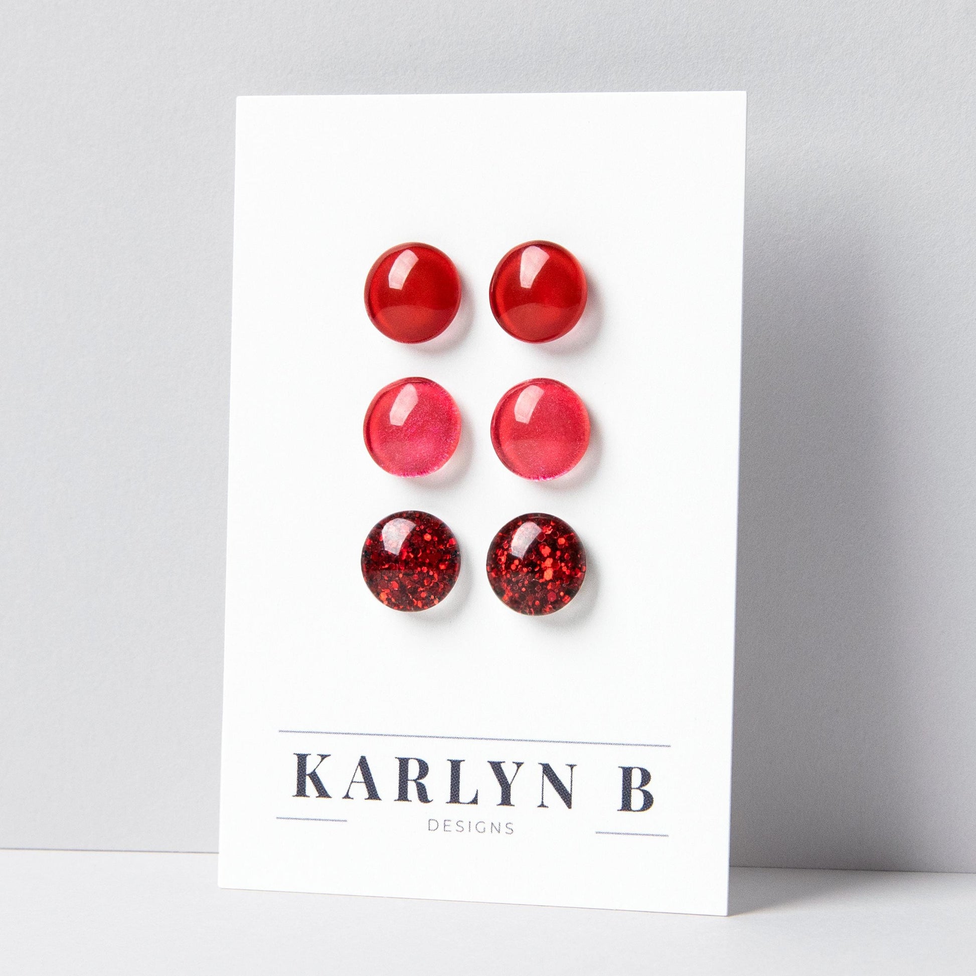 Set of three stud earrings in three different finishes - solid, shimmer, and glitter. The earrings are cherry red solid, ruby red shimmer, and ruby red glitter. The earrings are displayed on a white card with the text "KARLYN B DESIGNS".