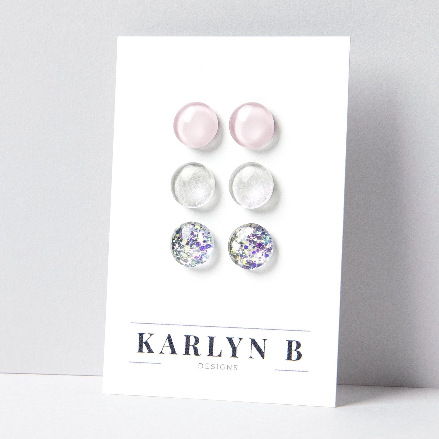 Set of three stud earrings in three different finishes - solid, shimmer, and glitter. The earrings are light pink, opal white shimmer, and opal silver glitter. The earrings are displayed on a white card with the text "KARLYN B DESIGNS".