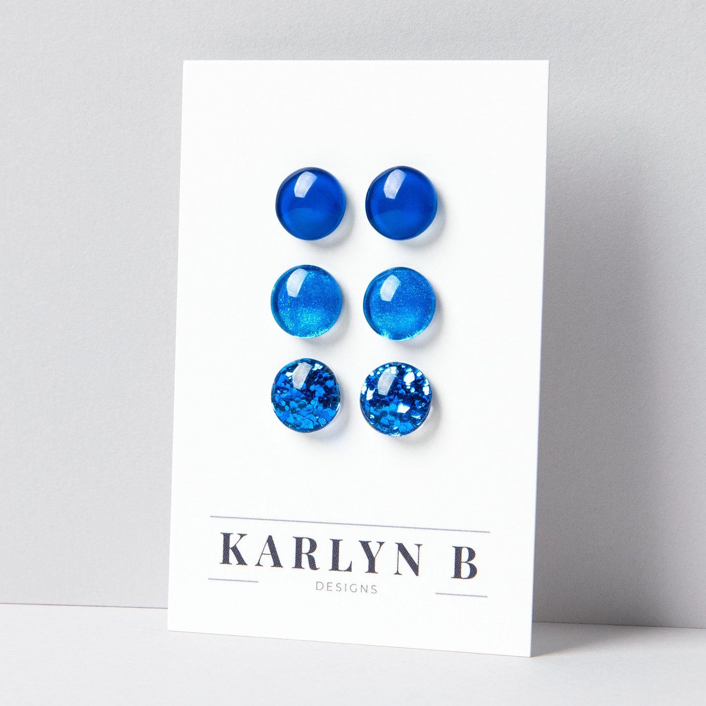 Set of three stud earrings in three different finishes - solid, shimmer, and glitter. The earrings are various shades of blue. The earrings are displayed on a white card with the text "KARLYN B DESIGNS".