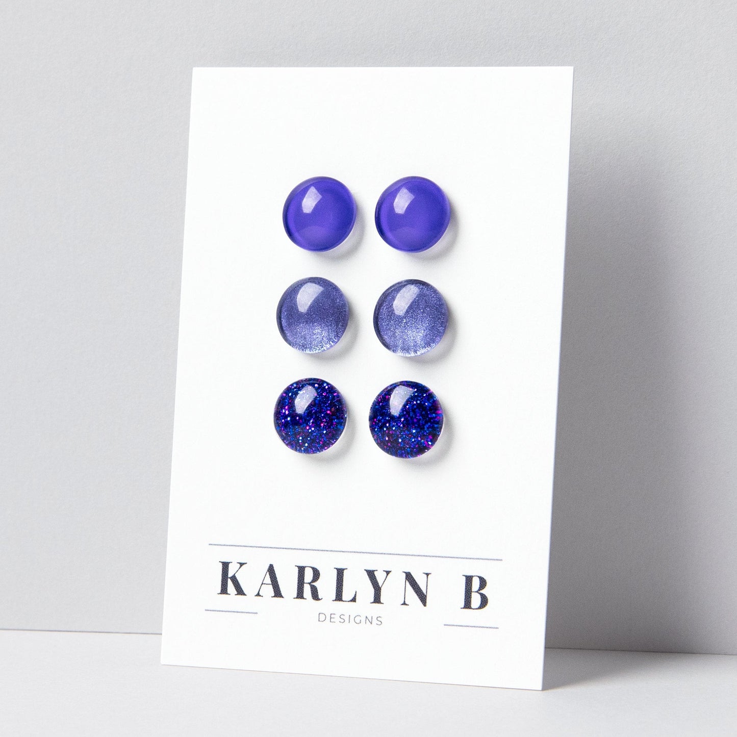 Set of three stud earrings in three different finishes - solid, shimmer, and glitter. The earrings are various shades of purple. The earrings are displayed on a white card with the text "KARLYN B DESIGNS".