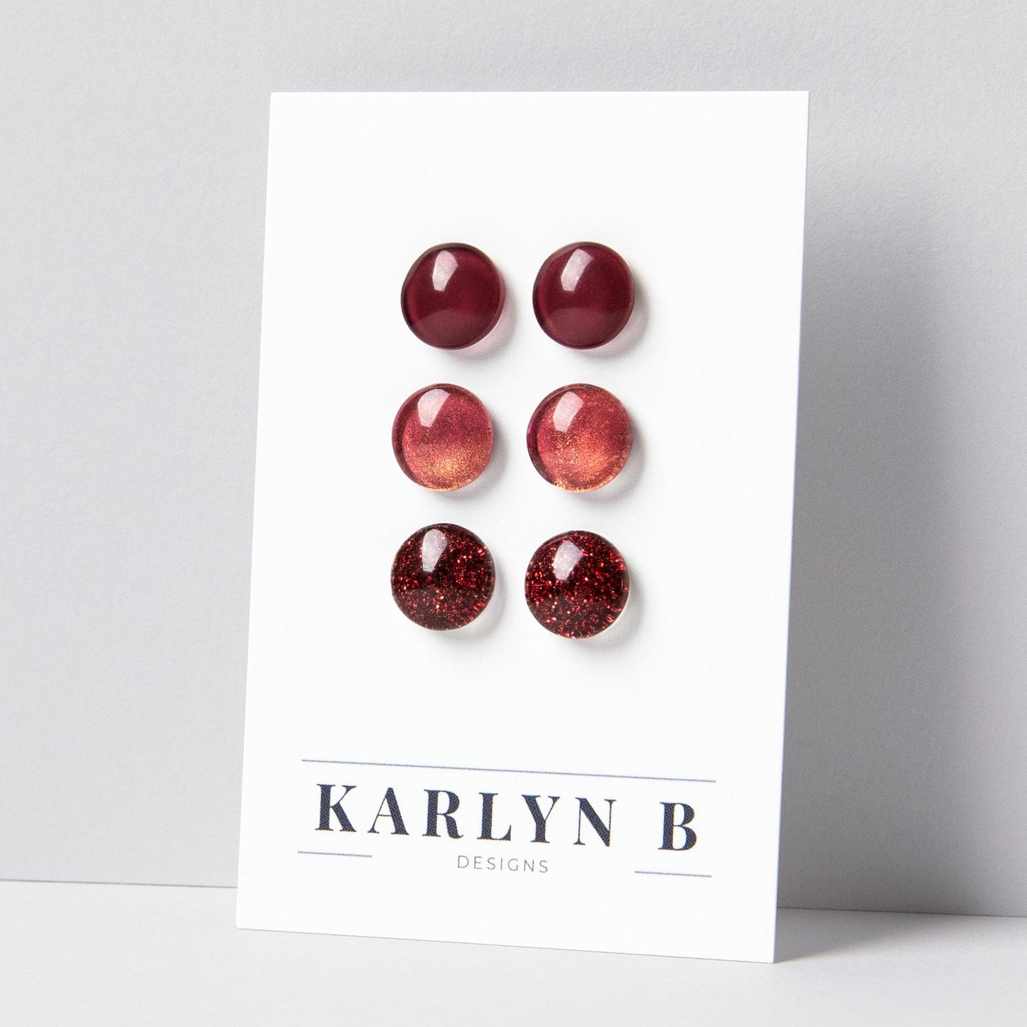 Set of three stud earrings in three different finishes - solid, shimmer, and glitter. The earrings are various shades of dark red. The earrings are displayed on a white card with the text "KARLYN B DESIGNS".