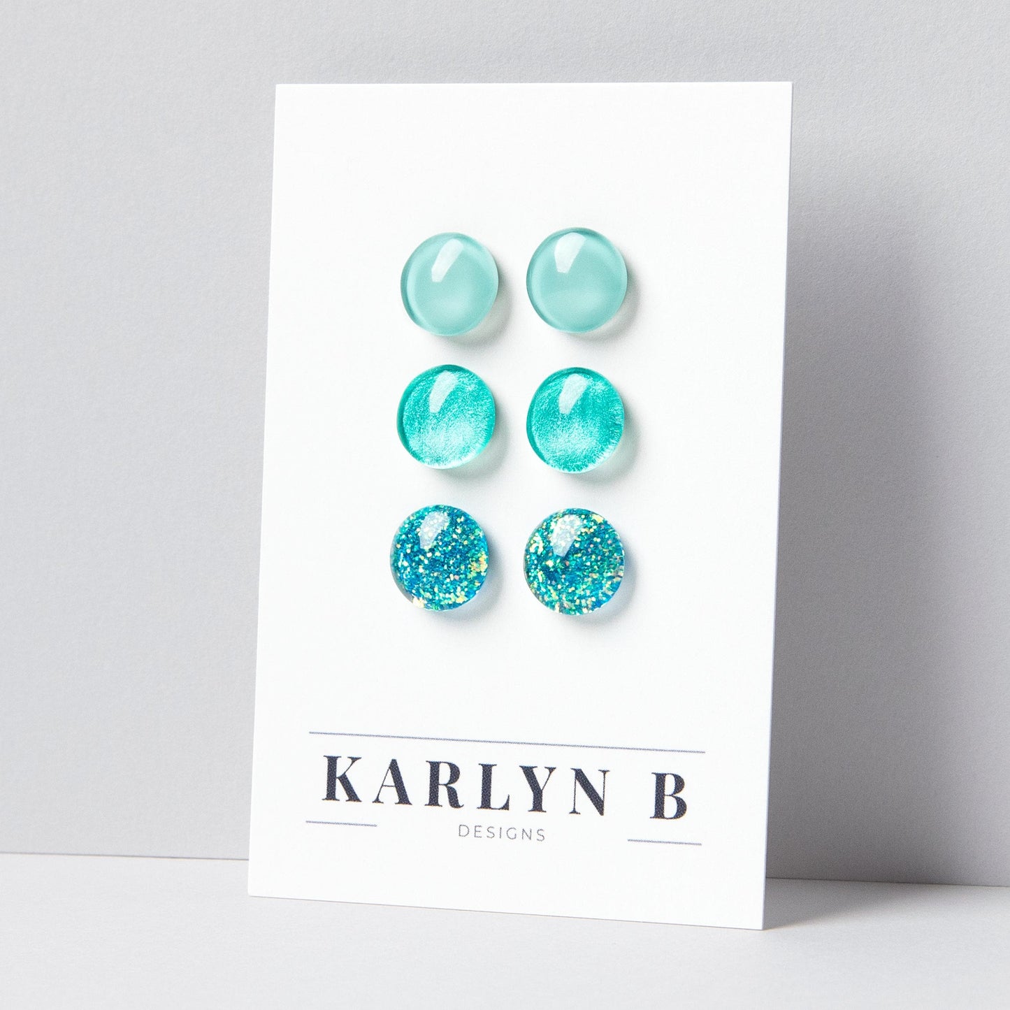 Set of three stud earrings in three different finishes - solid, shimmer, and glitter. The earrings are various shades of aquamarine. The earrings are displayed on a white card with the text "KARLYN B DESIGNS".
